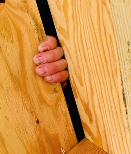 Inside an attic, fingers reaching through the open gap at the roof ridge between two boards.