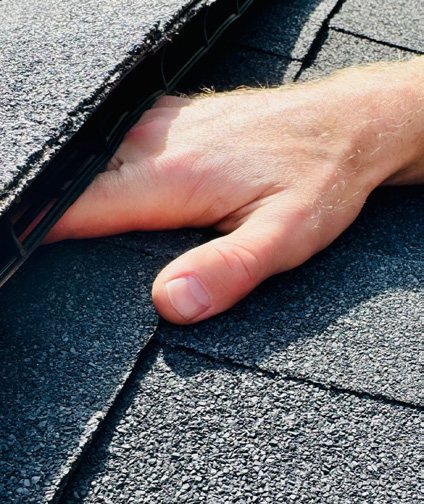 A hand sliding under a lifted ridge vent on a shingle roof, showing how wide the gap is.